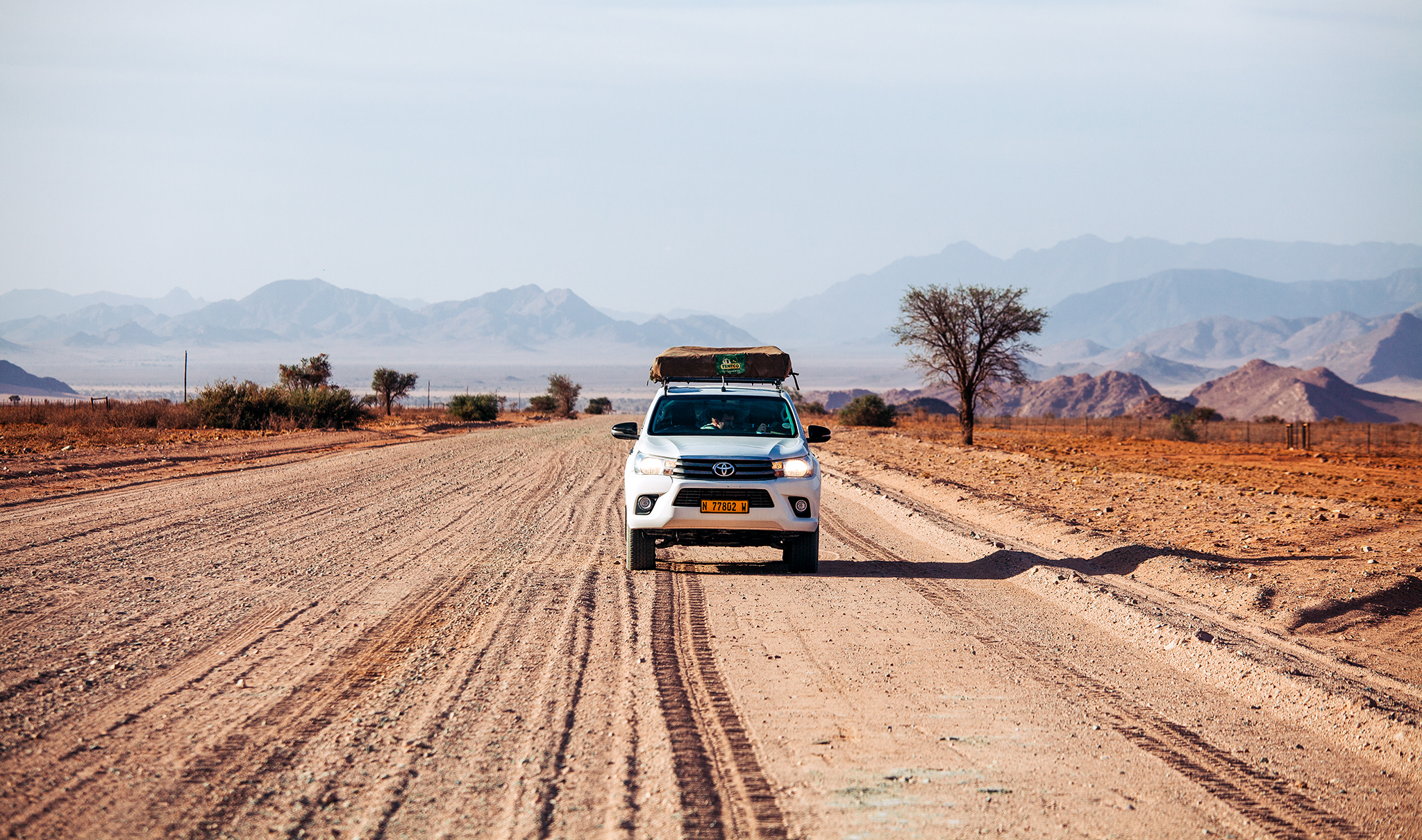 A white rental car with roof-top tent stands on a dirt road. Mountains in the background. Namibia in July is a great time for a self-drive safari. A white rental car with roof-top tent stands on a dirt road. Mountains in the background. Namibia in July is a great time for a self-drive safari.