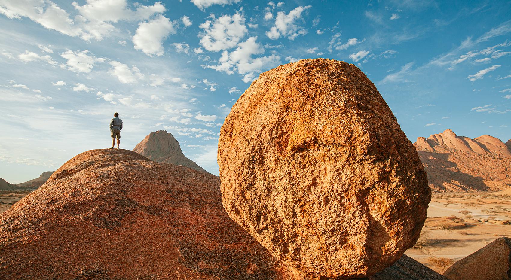 The landscape of Spitzkoppe. A man stands on a large rock. Mountains and blue sky in the background. The landscape of Spitzkoppe. A man stands on a large rock. Mountains and blue sky in the background.