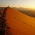 Two individuals standing on a large ochre-coloured sand dune at sunrise in Sossusvlei, Namibia.