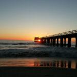 The sun setting into the Atlantic Ocean with the pier and waves crashing on the beach at Swakopmund are silhouetted.