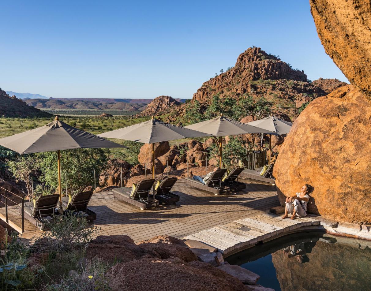A guest soaks up the sunshine, sitting next to a small pool, leaning against a large rock, while empty sun loungers and umbrellas overlook the stunning and otherworldly landscape of Damaraland.
